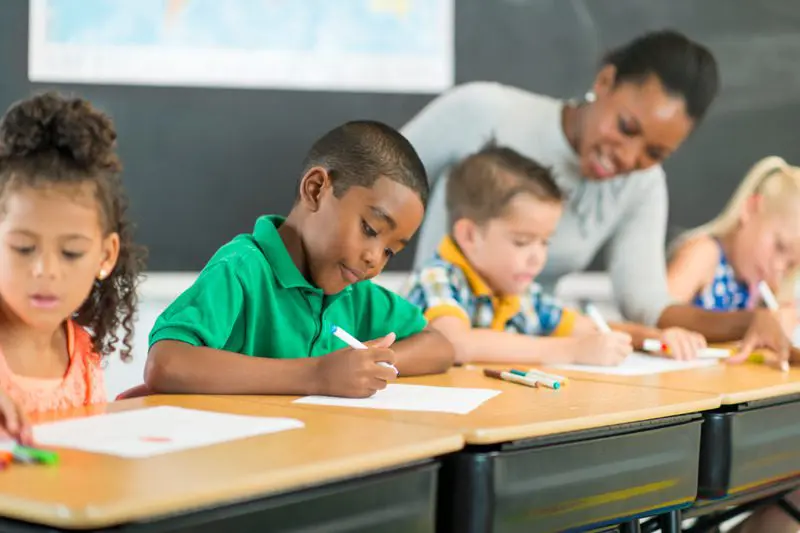 Students focusing on an assignment in a classroom while a teacher oversees their work.